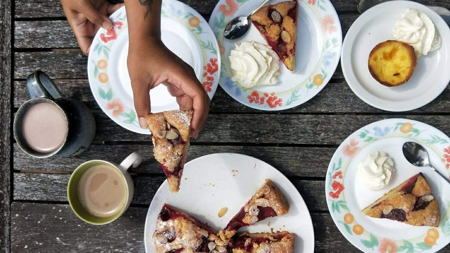 An outdoor table is laden with plates full of desserts. Pies with whipped cream. A hand reaches out to pass a piece of pie.
