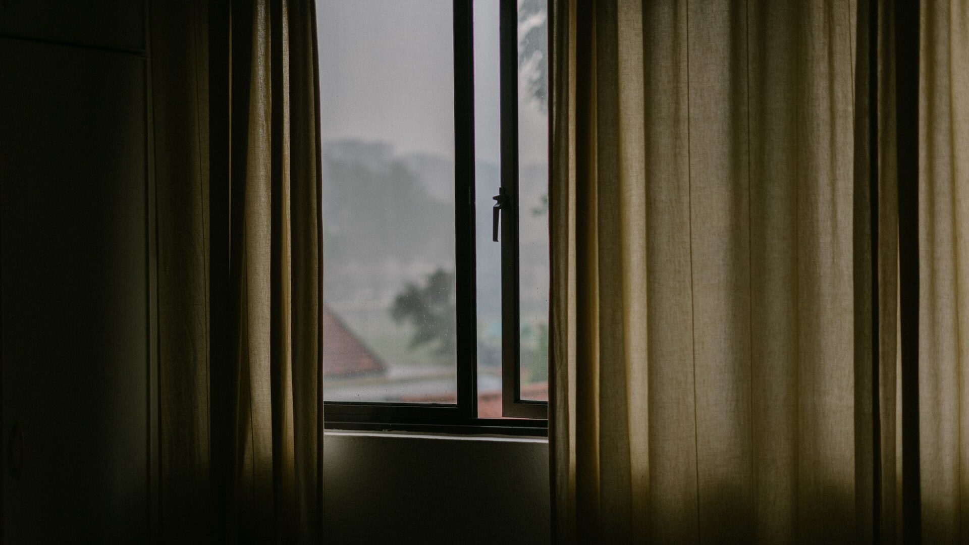 Half open curtains with an open window. It's a grey and rainy day outside.