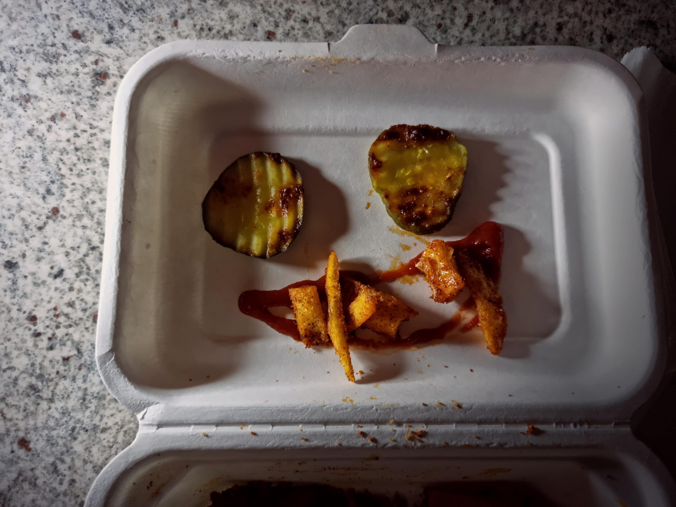 A smiley face made from pickles and fries lies in the top of a takeaway container.